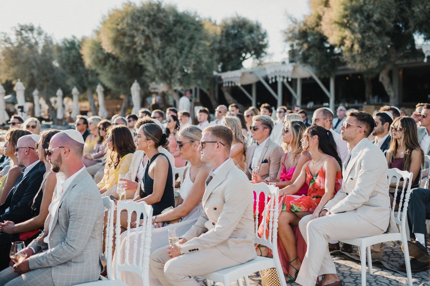 Outdoor wedding ceremony with guests seated in white chairs under trees.