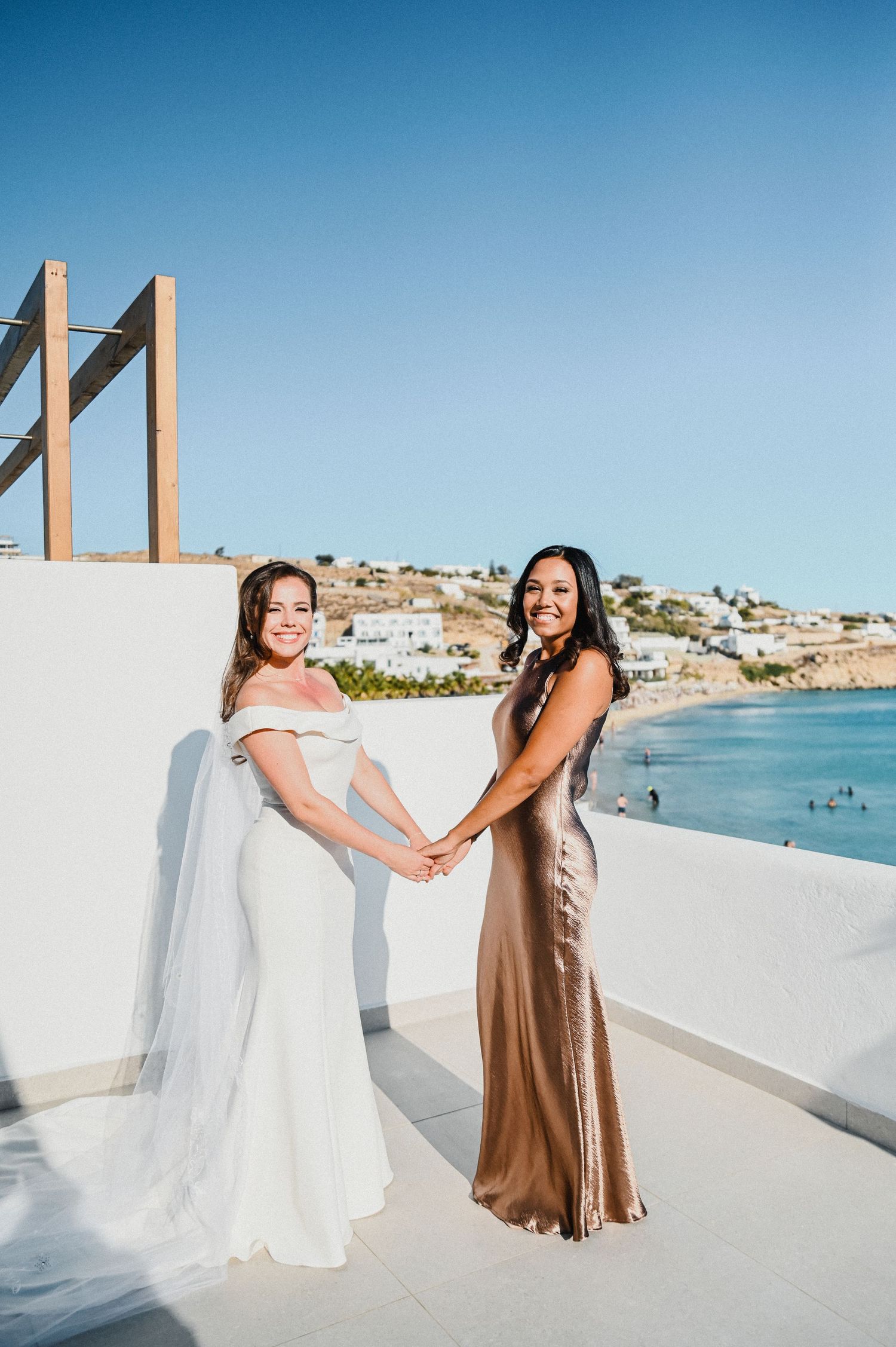 Two women hold hands on white terrace with coastal village backdrop.