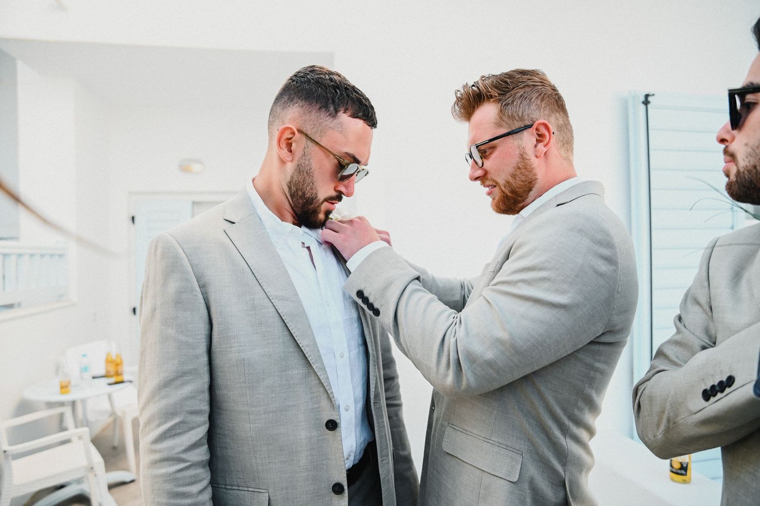 Two groomsmen in light gray suits help adjust wedding attire in an indoor setting.