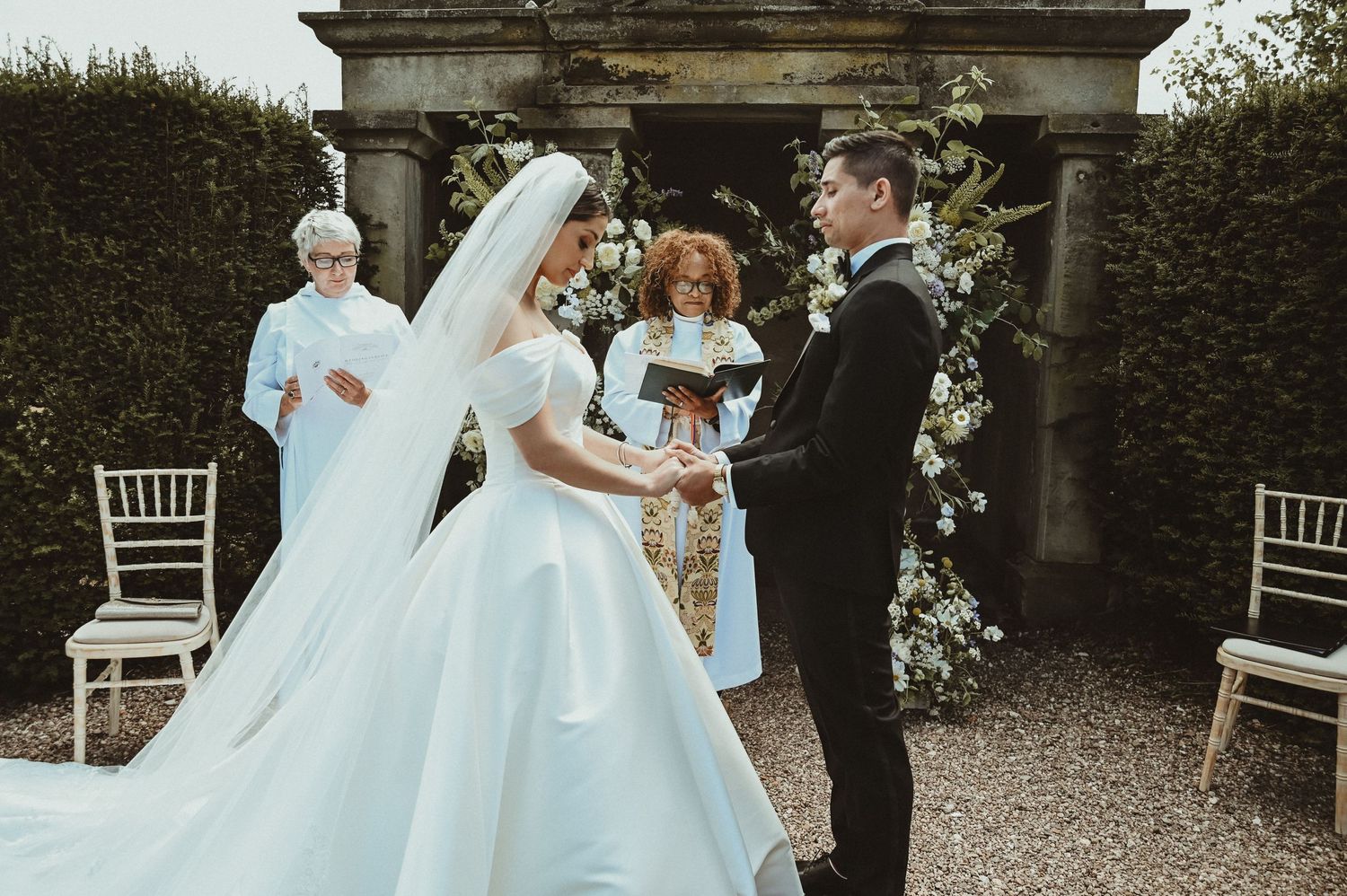 A wedding ceremony unfolds beneath an ornate stone archway adorned with white flowers.
