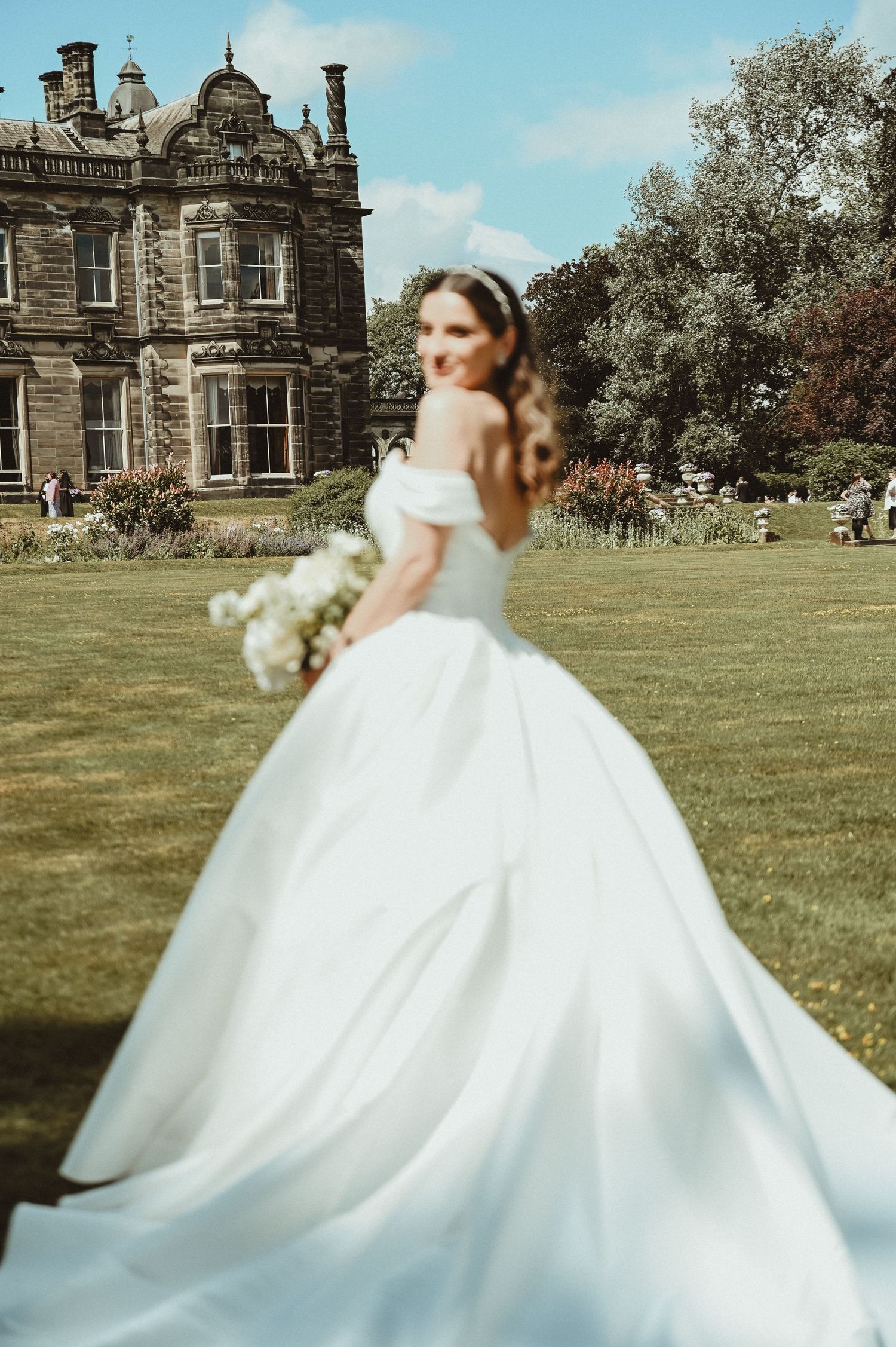 Elegant ballgown wedding dress flows across manicured lawn of grand Victorian mansion.
