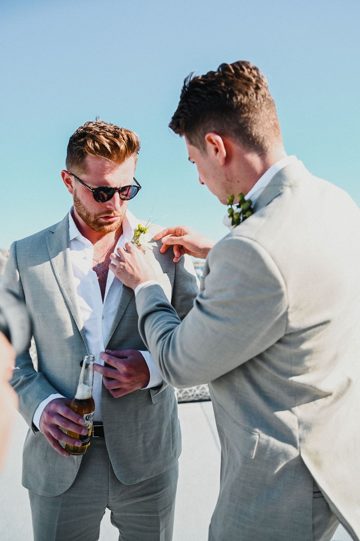 Two men in gray suits adjusting boutonnieres against blue sky background.