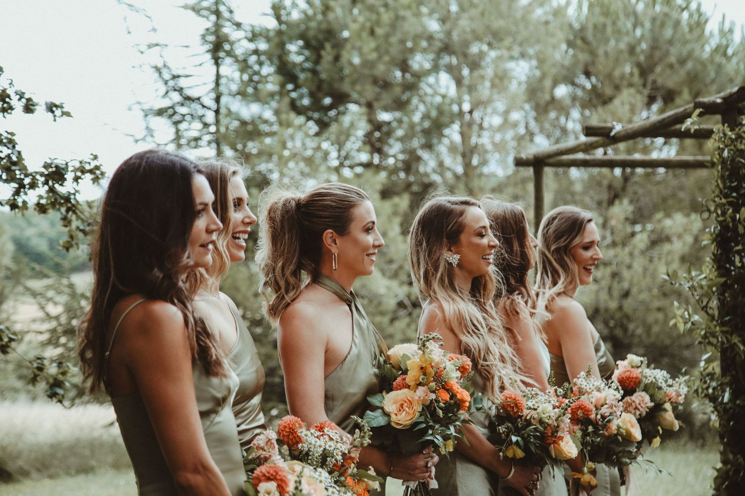 Bridesmaids in sage green dresses hold colorful autumn bouquets during an outdoor ceremony.