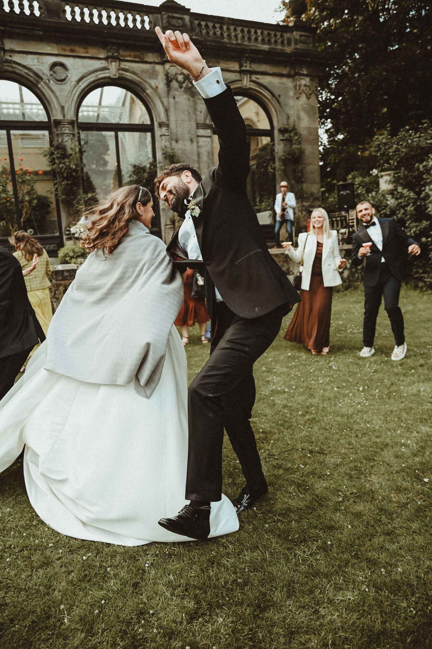 Wedding celebration with dancing couples on a lawn in front of an ornate stone archway.