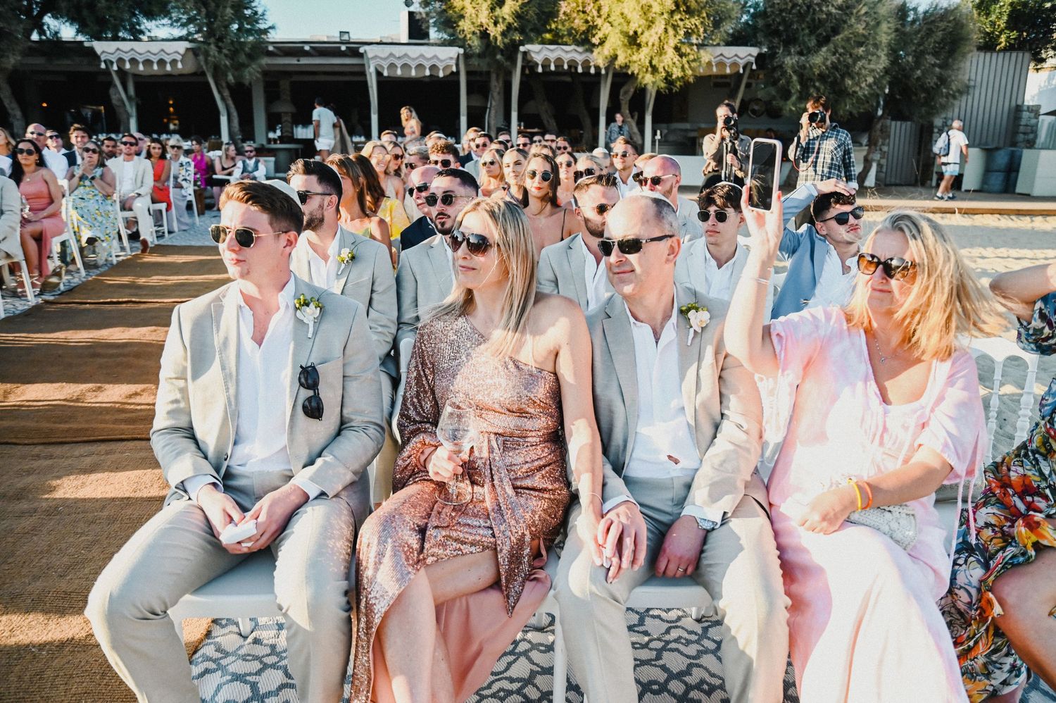 Wedding guests in summer attire seated outdoors with sunglasses at beach venue.