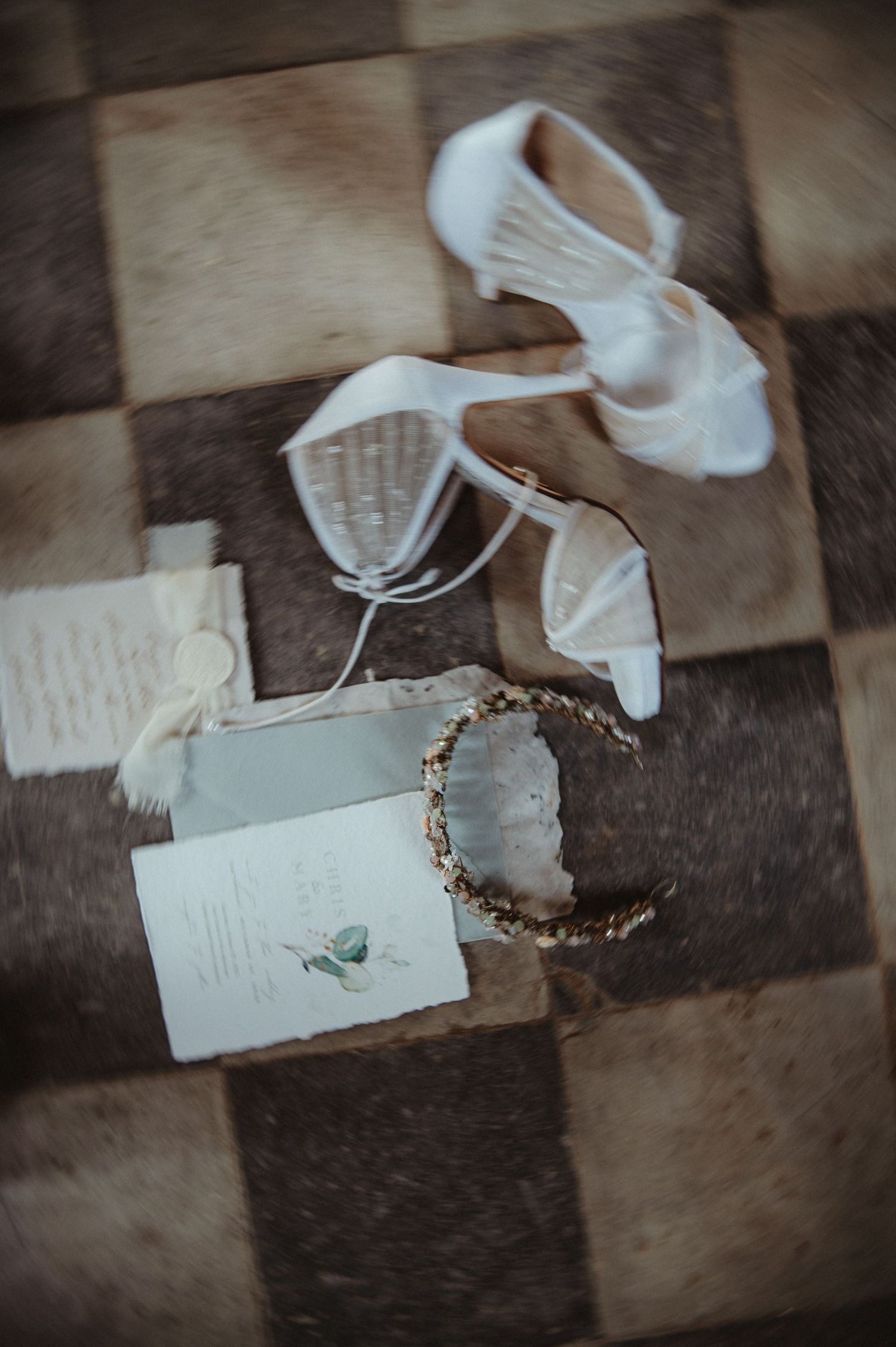 Wedding accessories including white shoes, invitation and delicate crown on checkerboard floor.
