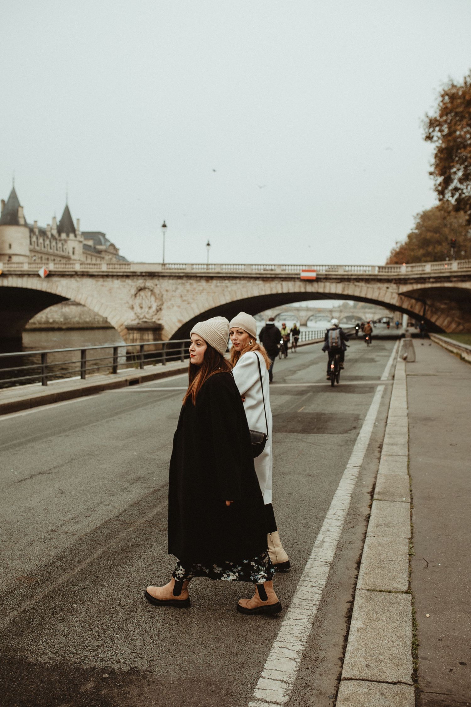 Two people in warm coats standing along the Seine River in Paris with a historic bridge and architecture in the background.