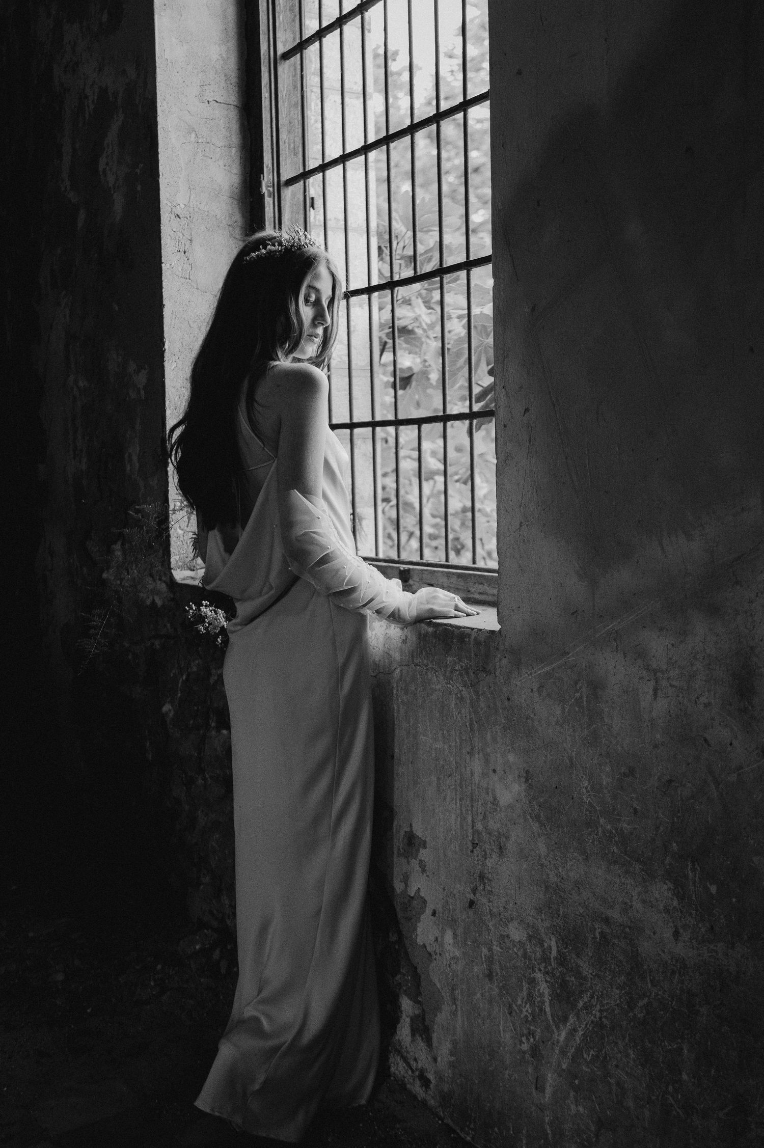 Elegant silhouette of bride in form-fitting dress gazing through industrial window.