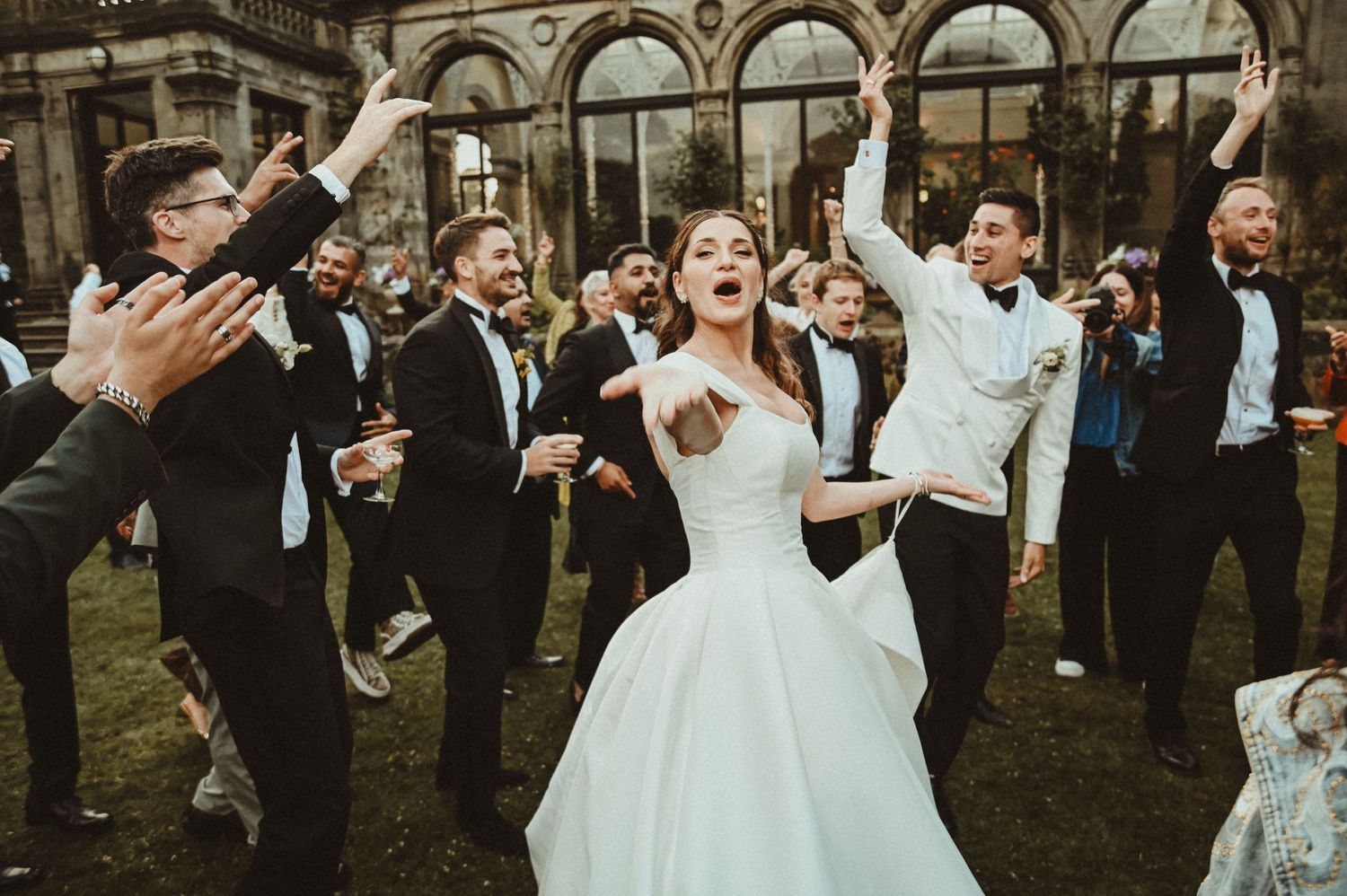 Wedding party celebrates with raised arms and enthusiastic dancing outdoors.
