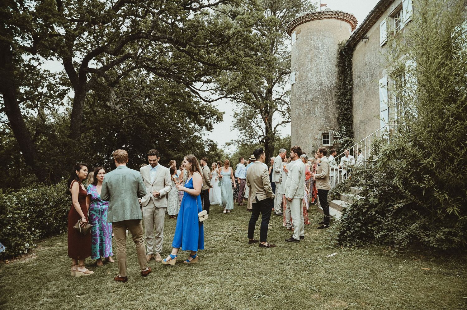 Wedding guests gather on lawn outside historic chateau during outdoor reception.