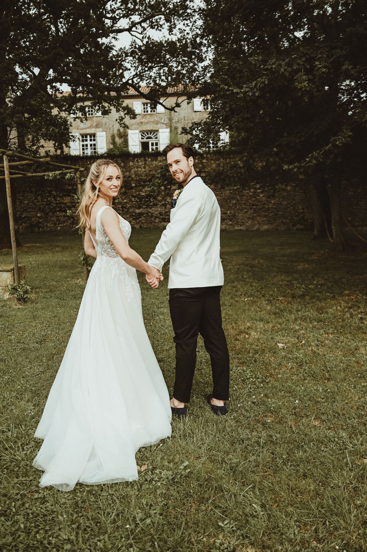 A wedding couple poses in front of a grand stone house on a lush green lawn.