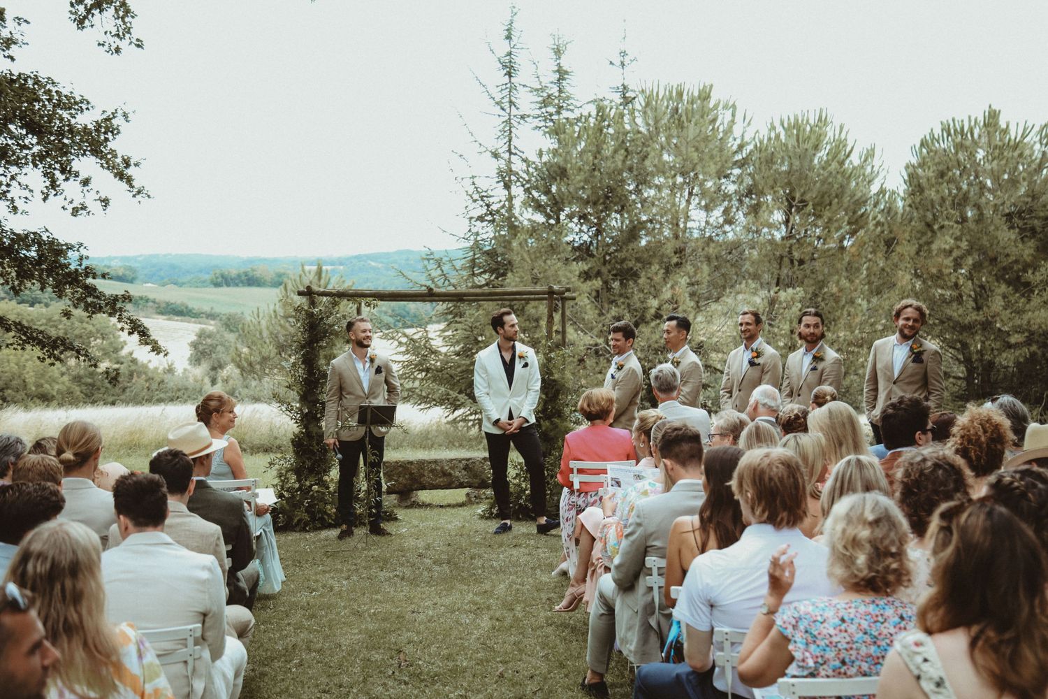 Outdoor wedding ceremony with guests seated in white chairs surrounded by trees and natural scenery.