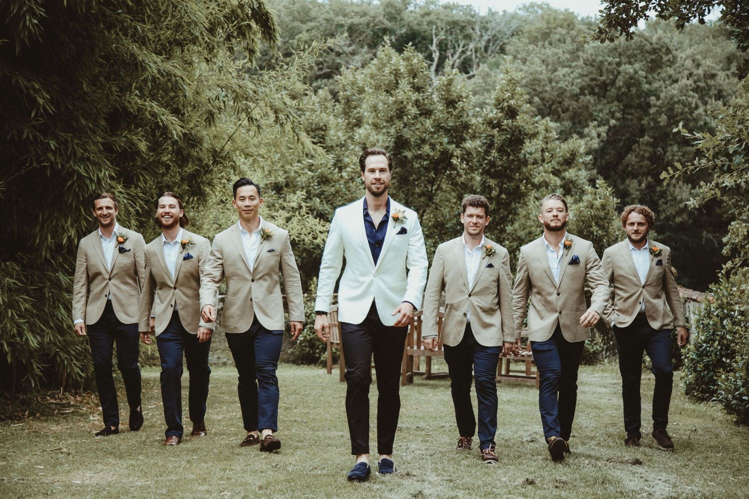 Groomsmen in beige suits follow person in white suit walking through garden setting.