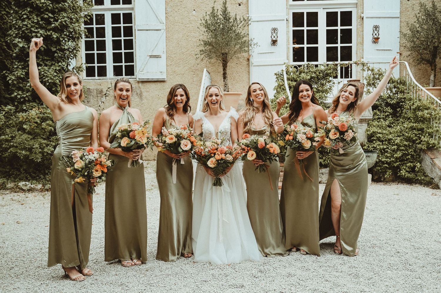 Wedding party celebrating with raised arms and floral bouquets in front of a classic building exterior.