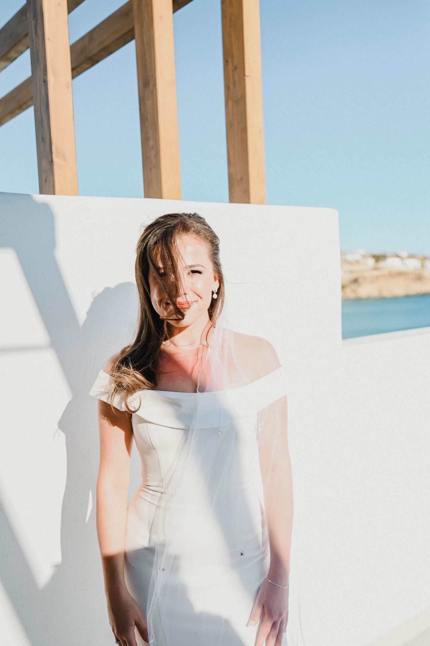 Sunlit portrait of bride in white dress against white wall and wooden beams.