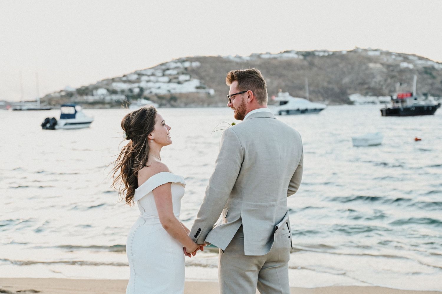 Newlyweds share intimate moment while holding hands on beach with boats in background.