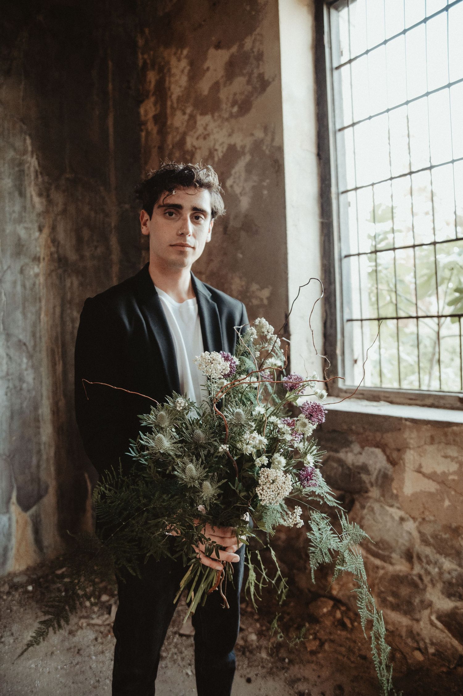 Moody portrait with person in dark suit holding rustic wildflower bouquet by window.