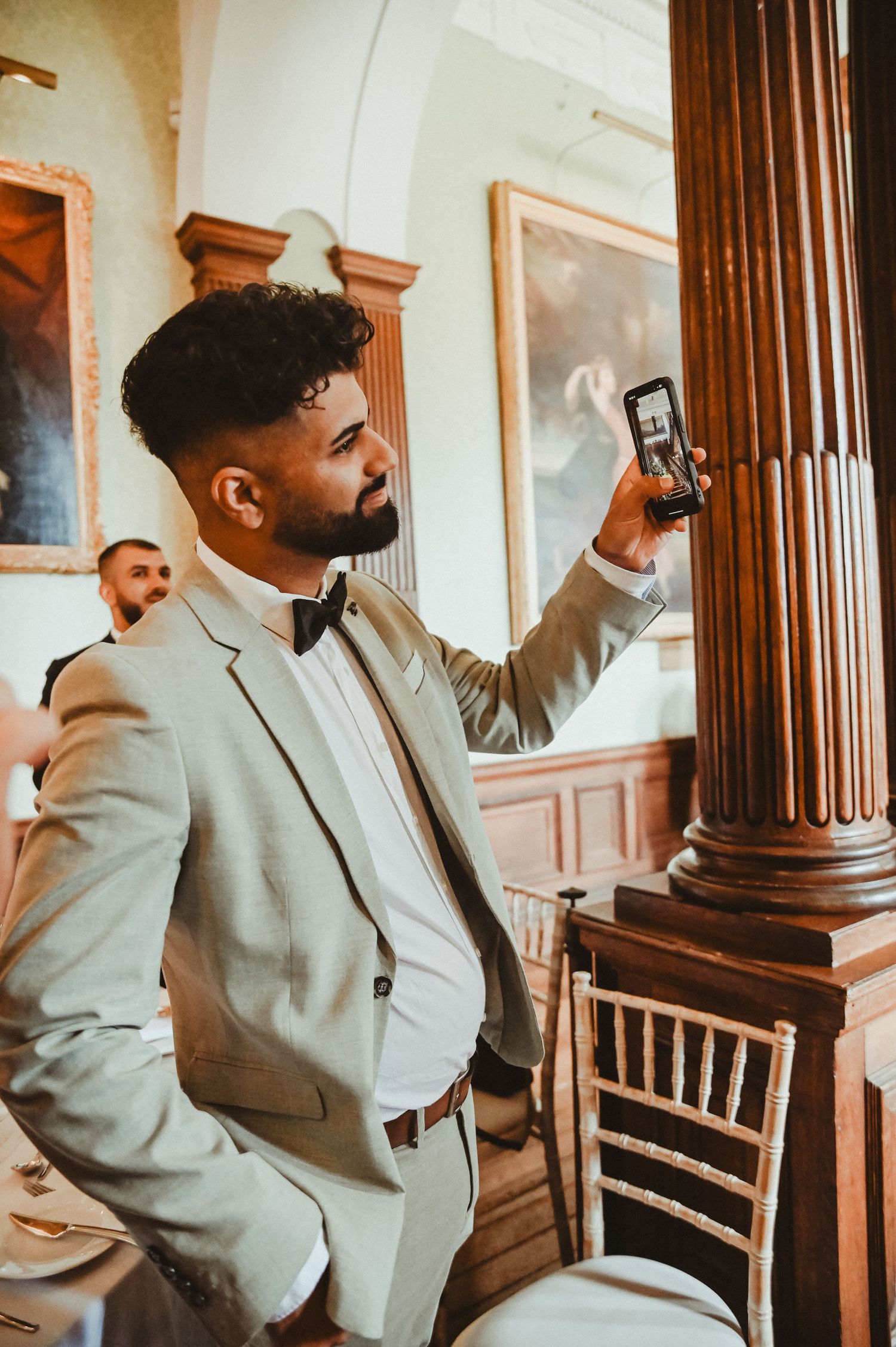 Wedding guest in light gray suit taking a photo with smartphone in an ornate interior hallway.