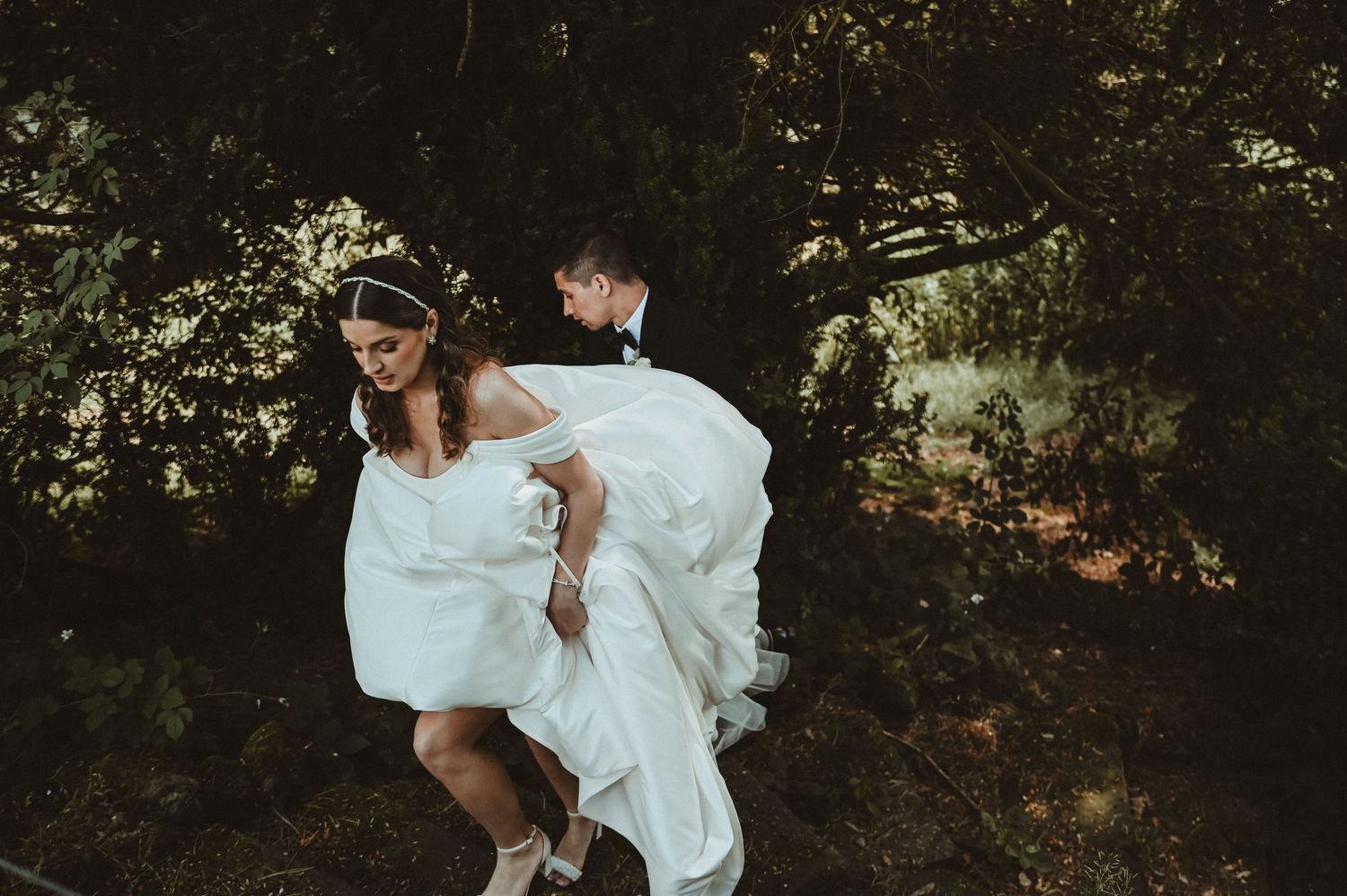 Couple embraces in woodland setting with flowing white dress creating romantic atmosphere.
