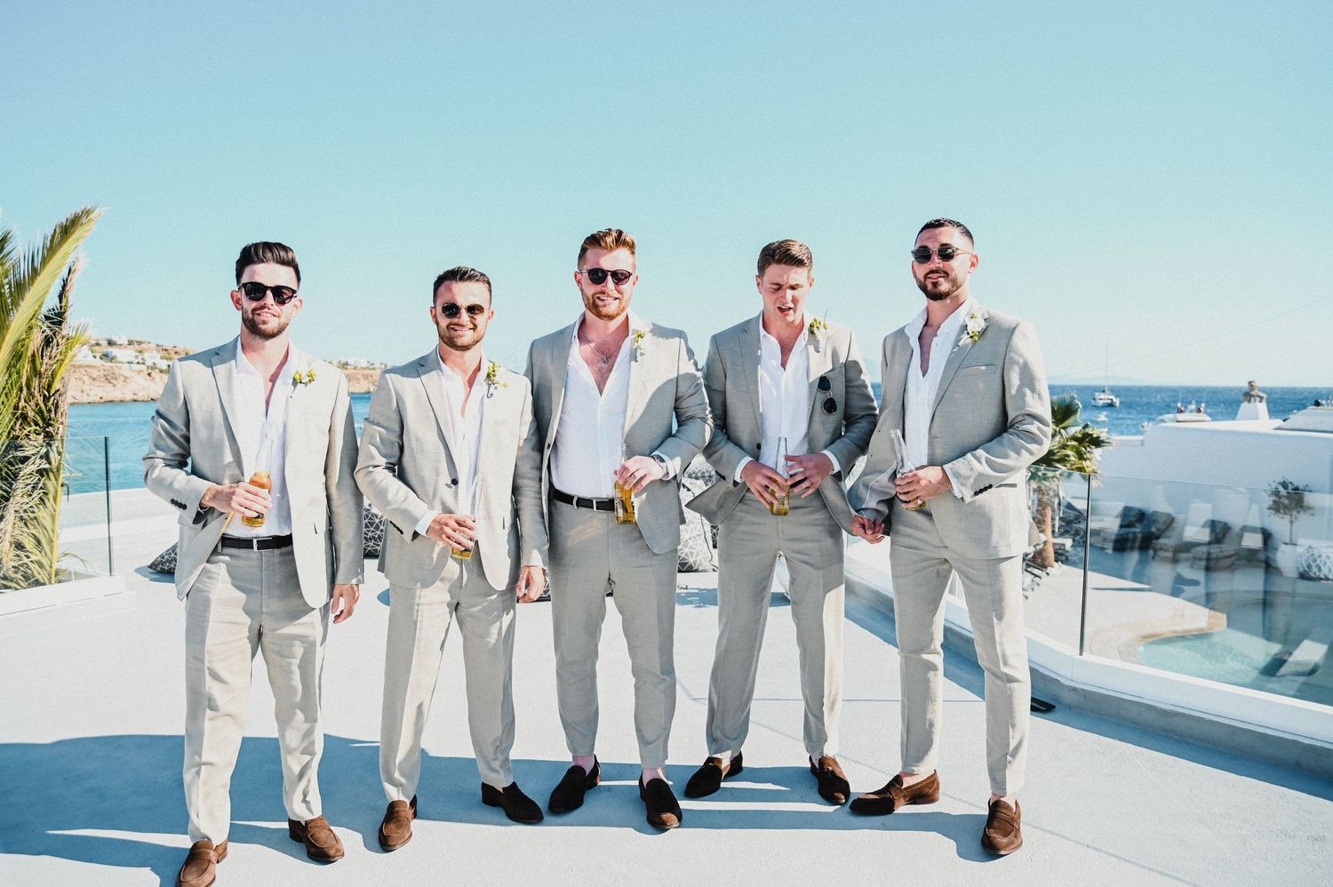 Wedding party in matching light gray suits pose on Mediterranean terrace with ocean view.