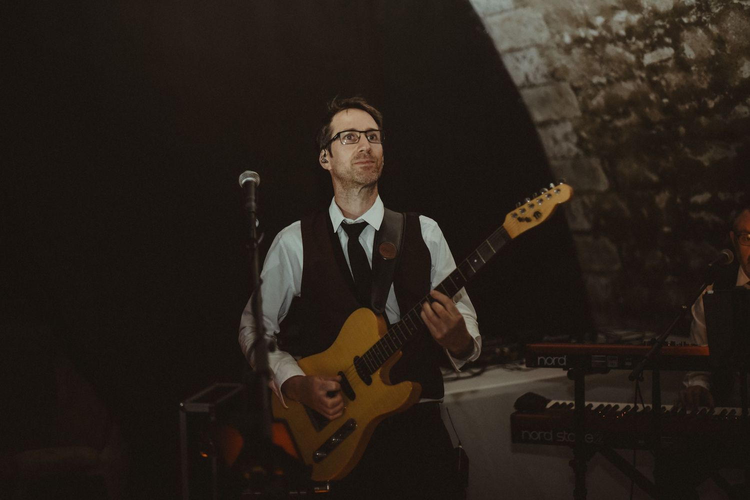 A musician plays electric bass guitar while performing on stage in a dimly lit venue.