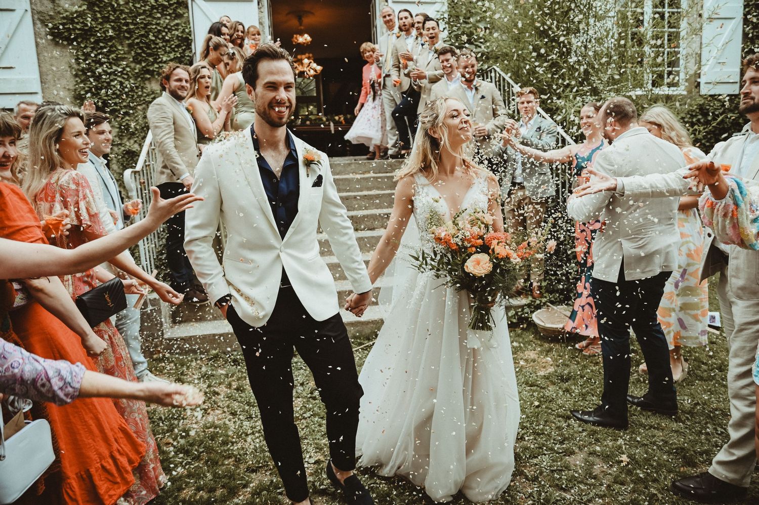 Wedding celebration with guests in formal attire throwing confetti outside stone building.