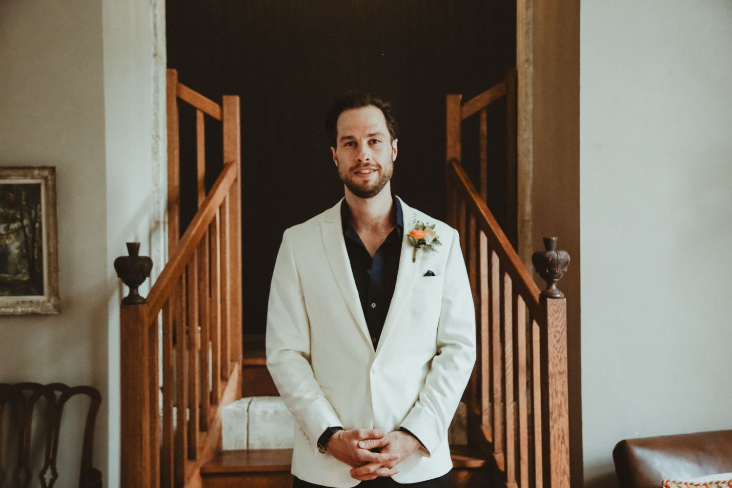 Groom in white blazer and dark shirt poses in front of wooden staircase.