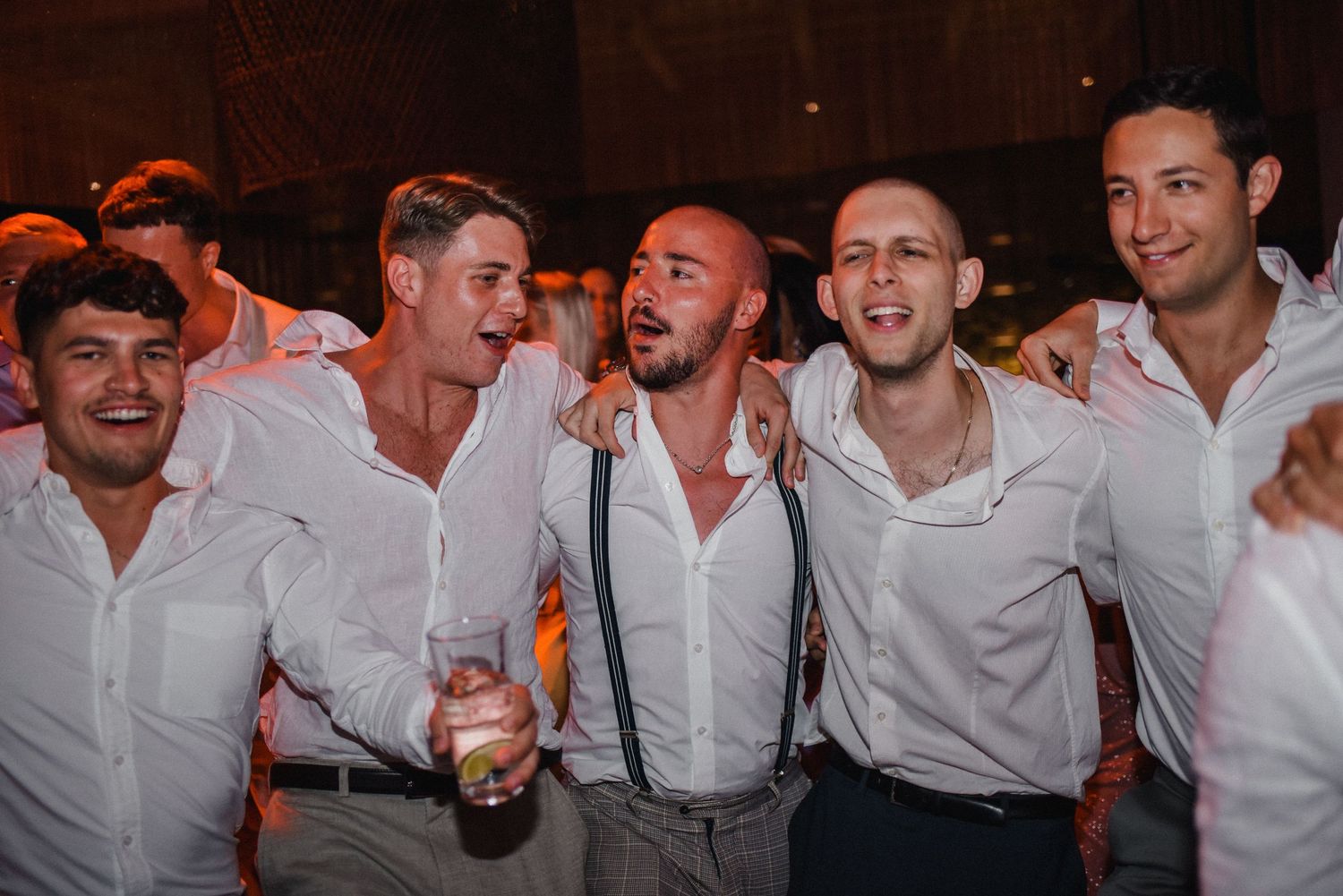 Group of groomsmen in white shirts and suspenders celebrating at wedding reception.