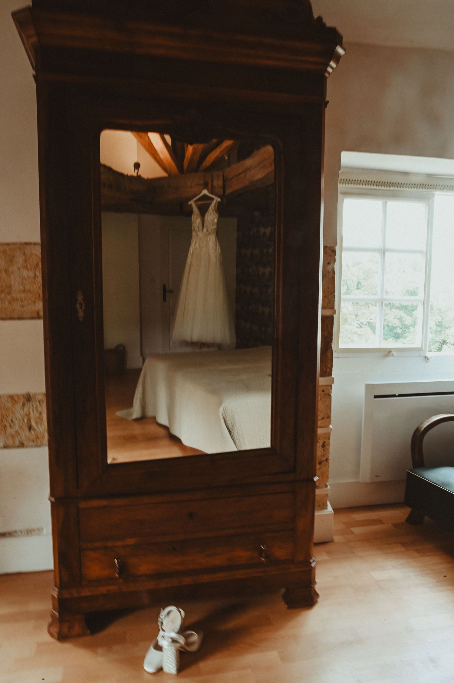 White wedding dress hangs elegantly on a vintage wooden armoire with mirror reflection.