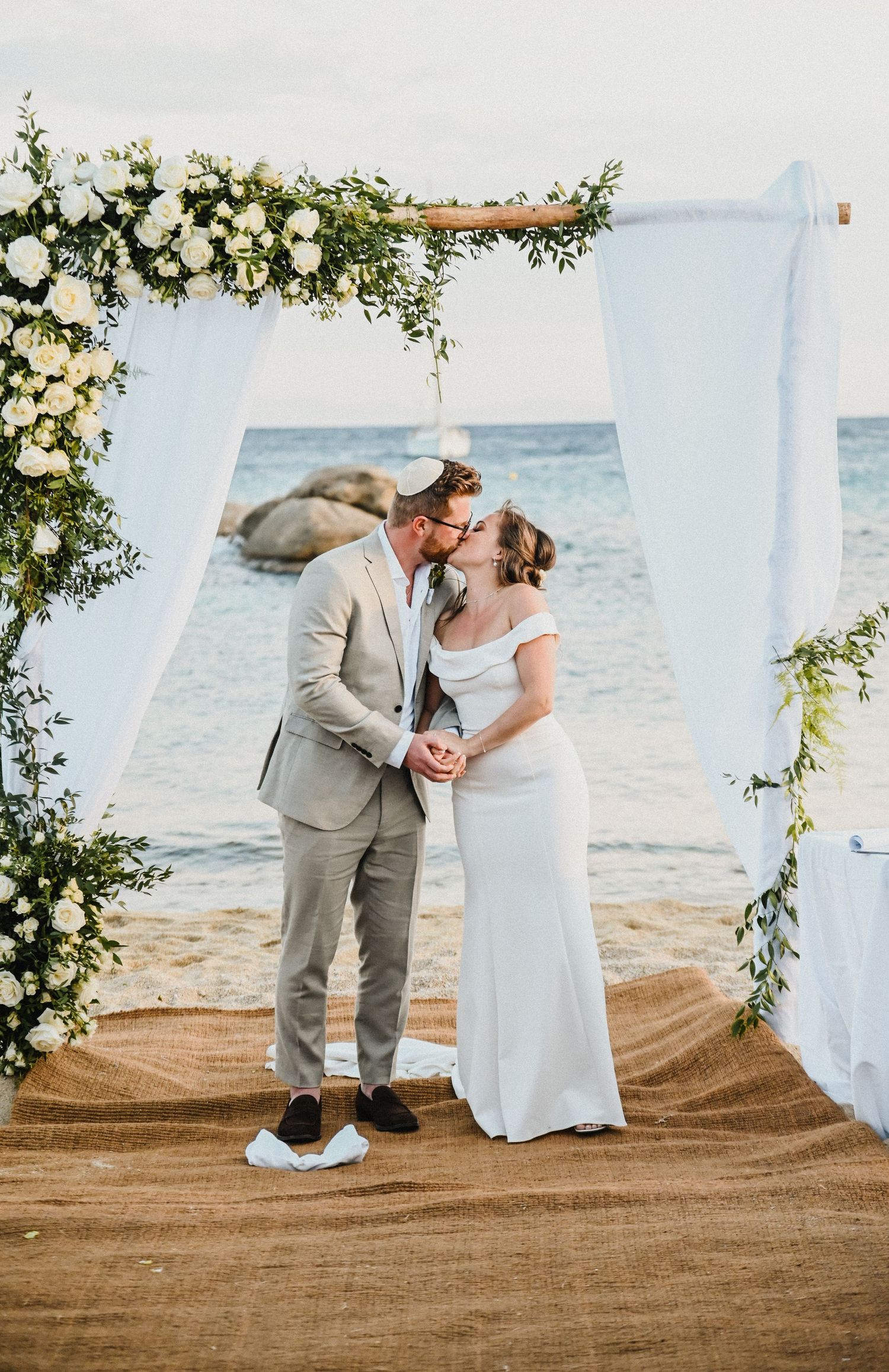 Couple shares romantic moment under floral arch during beach wedding ceremony.