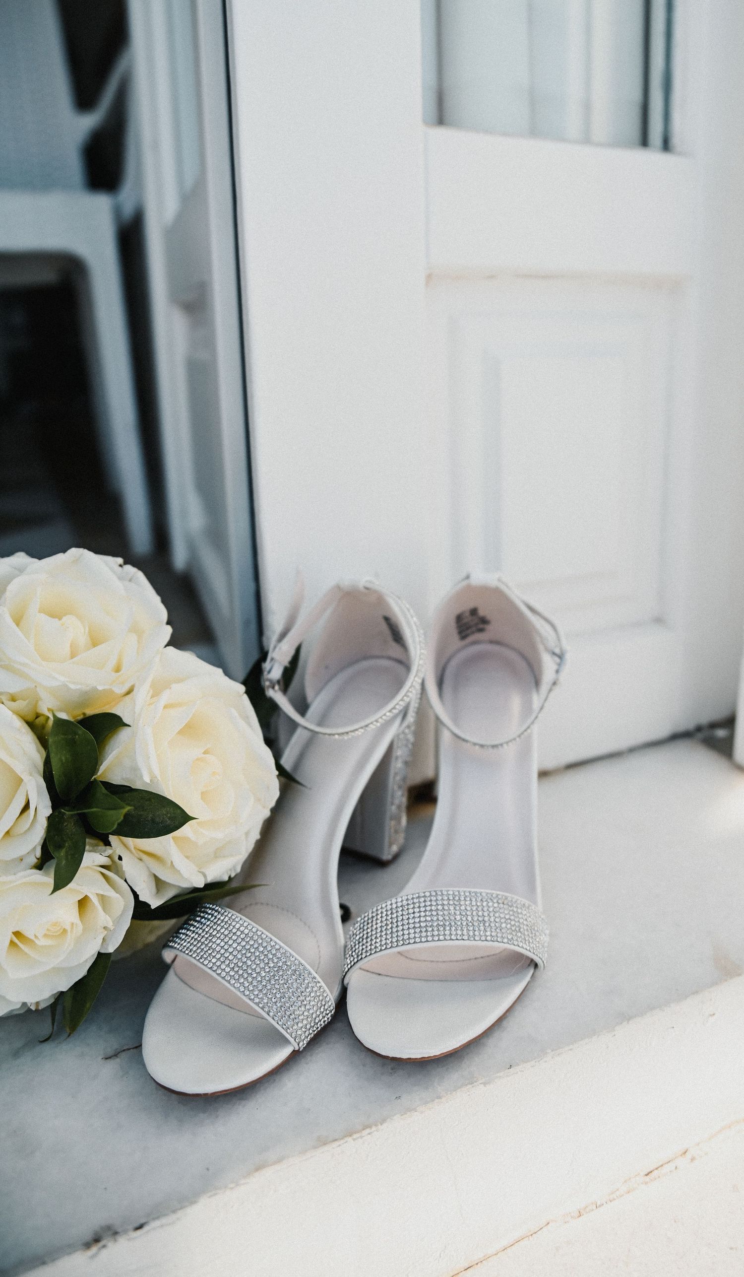 Elegant bridal shoes with rhinestone details placed next to white roses on a windowsill.