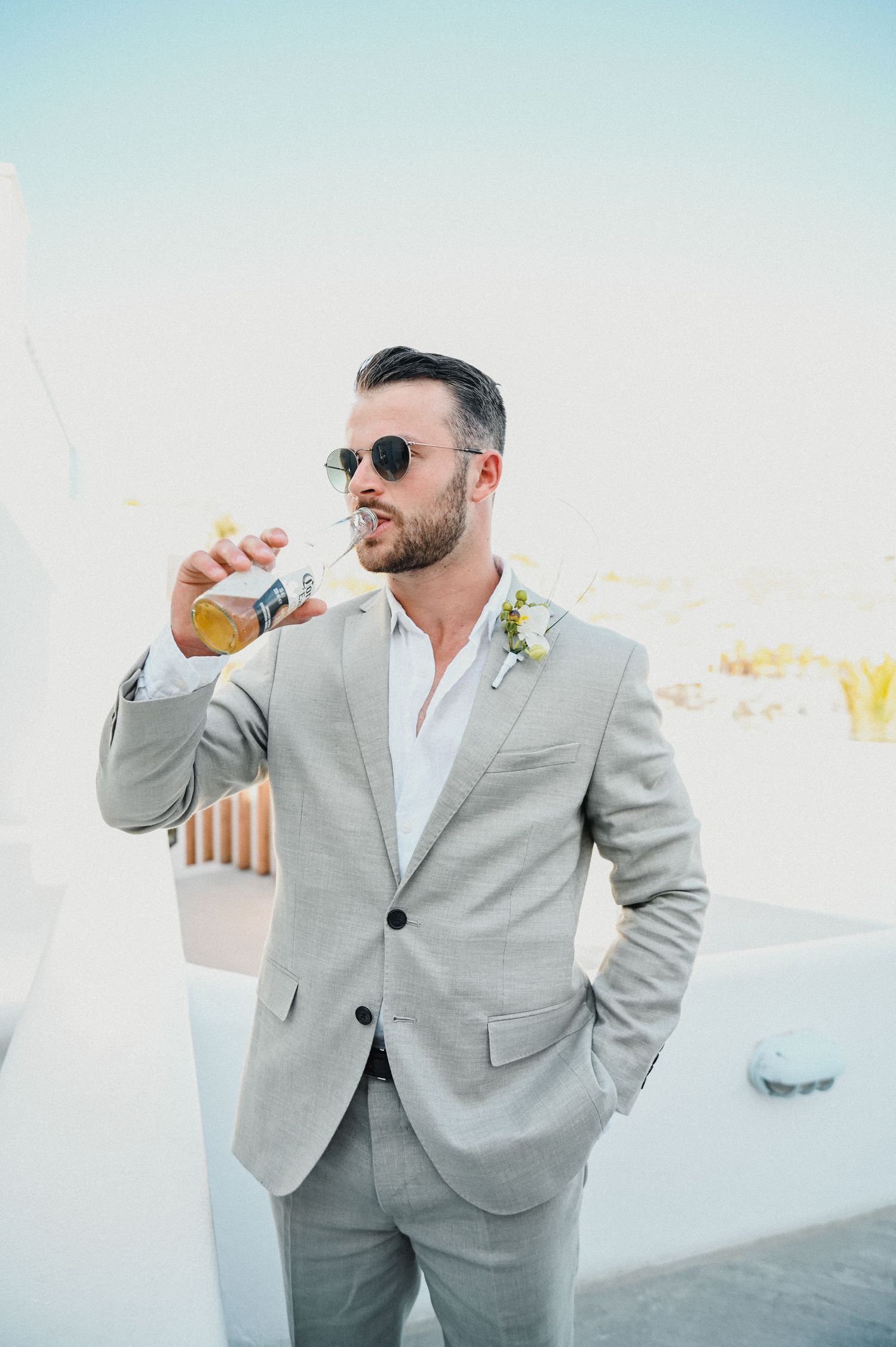 Stylish groomsman in light gray suit and sunglasses drinks from a bottle on a white terrace.