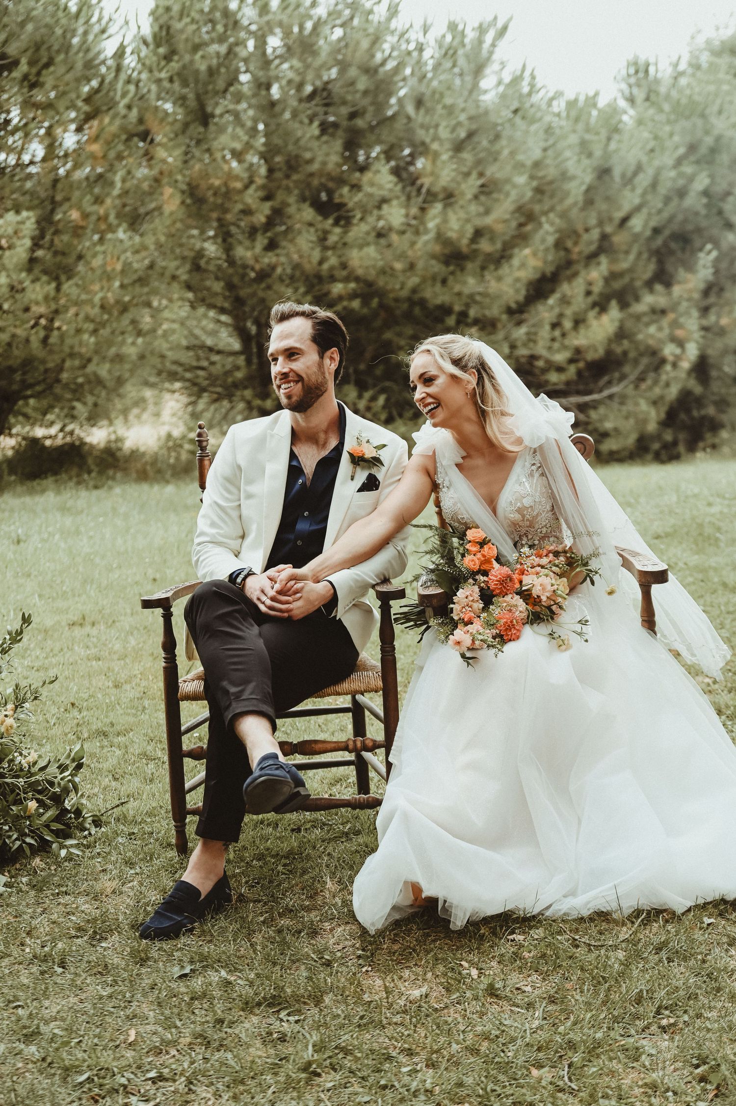 Newlyweds share a joyful moment on rustic chairs during their outdoor garden celebration.