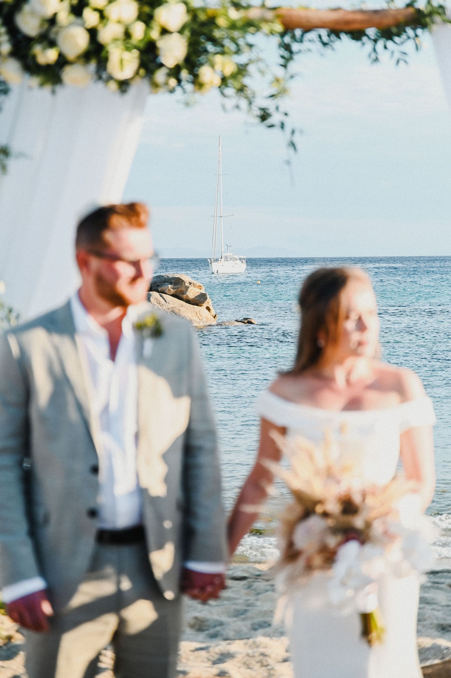 Beachside wedding ceremony with sailboat visible in background through floral arch.