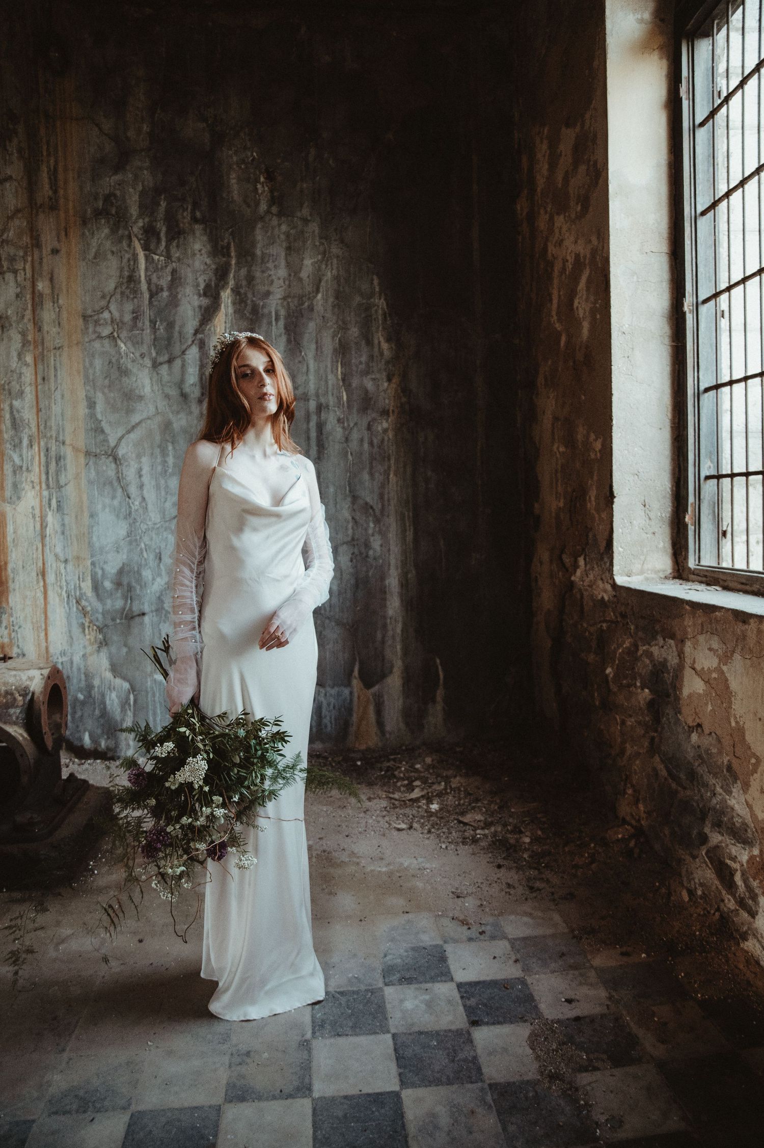 Ethereal bridal portrait in flowing white dress standing by window in abandoned building with checkerboard floor.