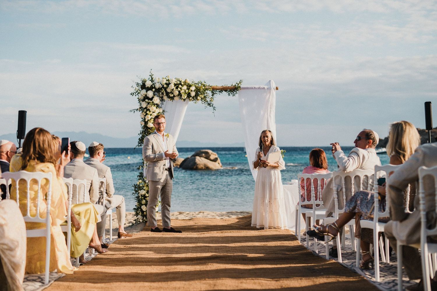 Outdoor beach wedding ceremony with white chairs and floral arch against ocean backdrop.
