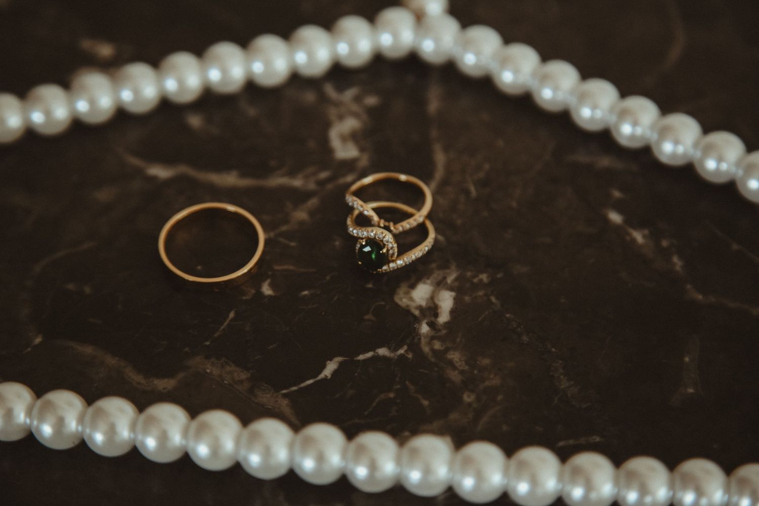 Pearl necklace and engagement rings arranged on dark marble surface.