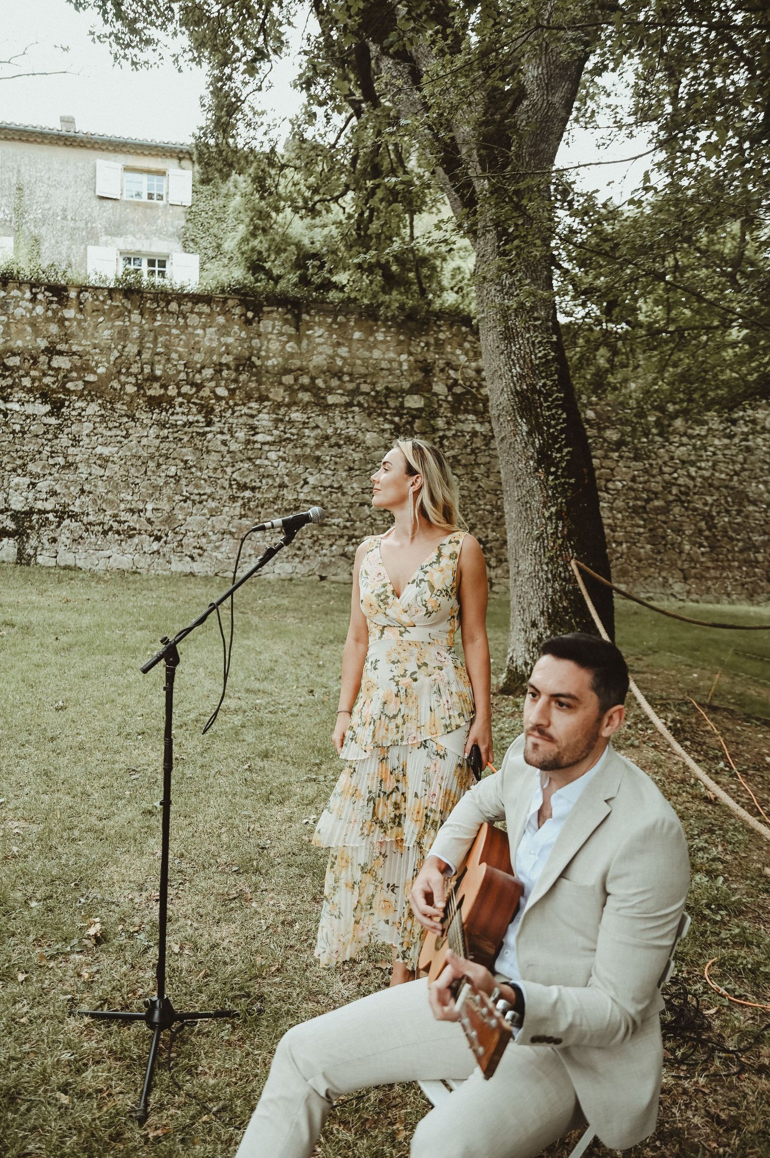 Musicians performing at outdoor wedding with stone wall and tree backdrop.