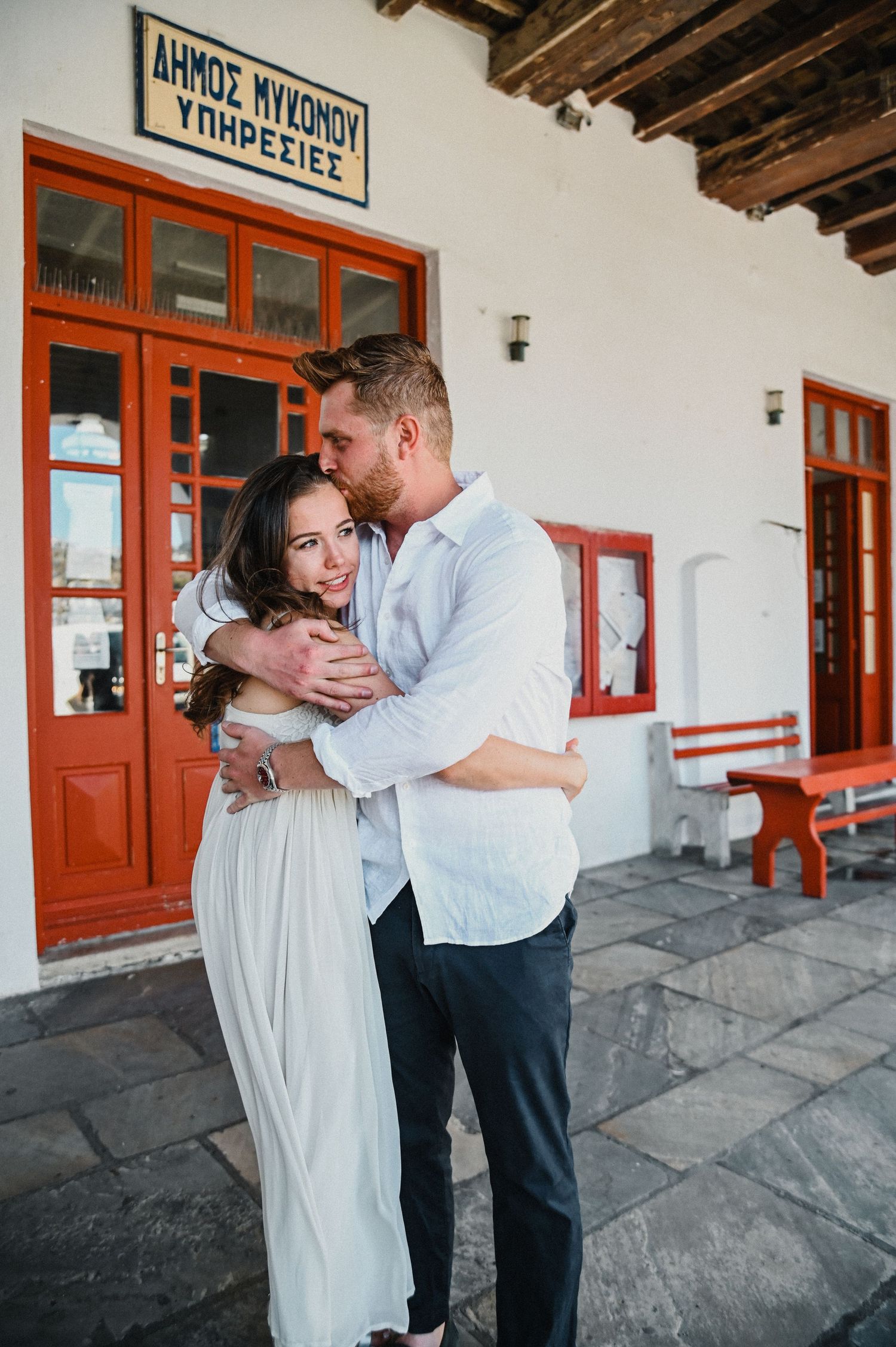 Romantic embrace outside a traditional Greek building with red doors and a rustic sign.