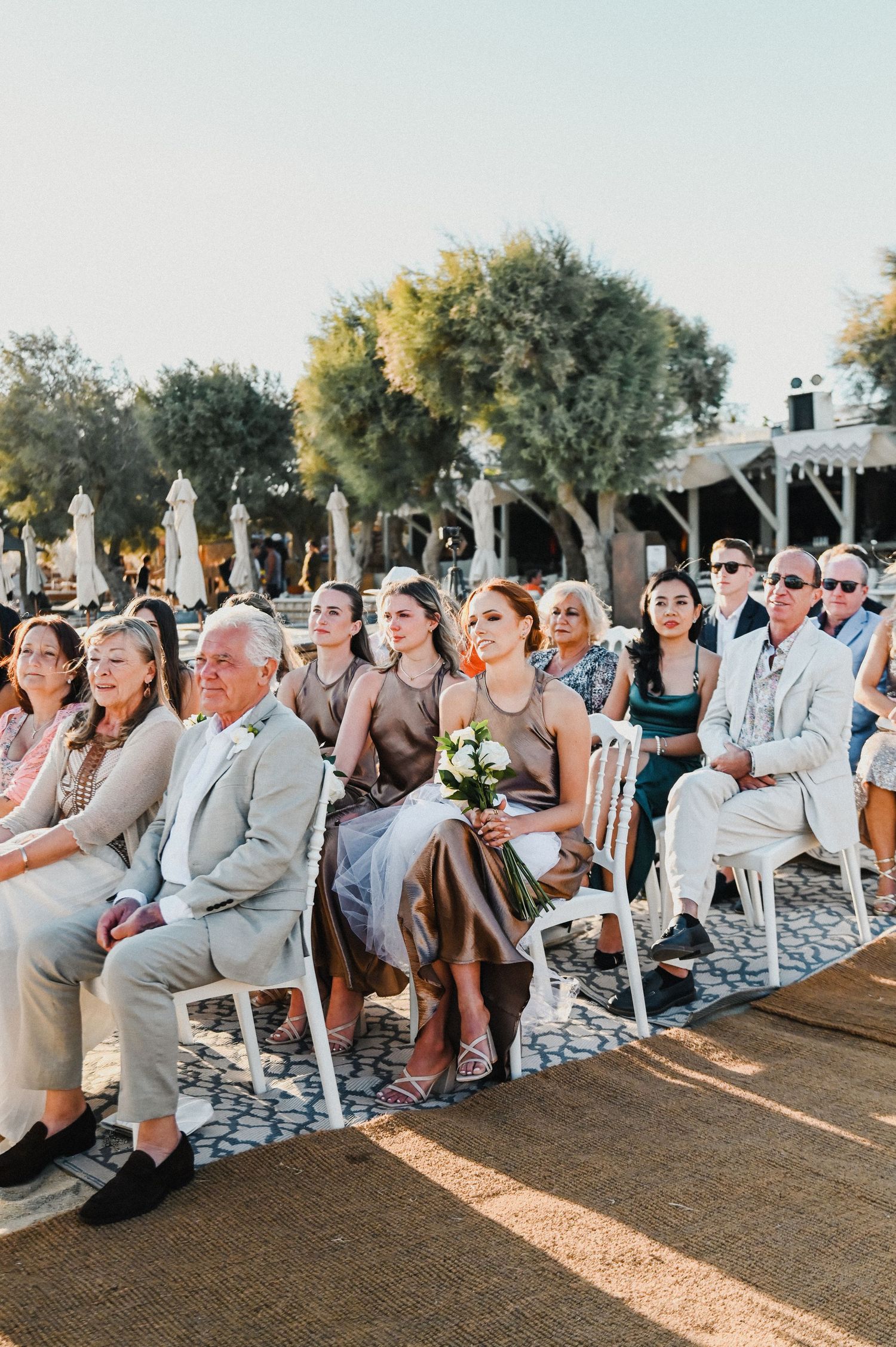 Wedding guests seated in rows during outdoor ceremony under trees at sunset.