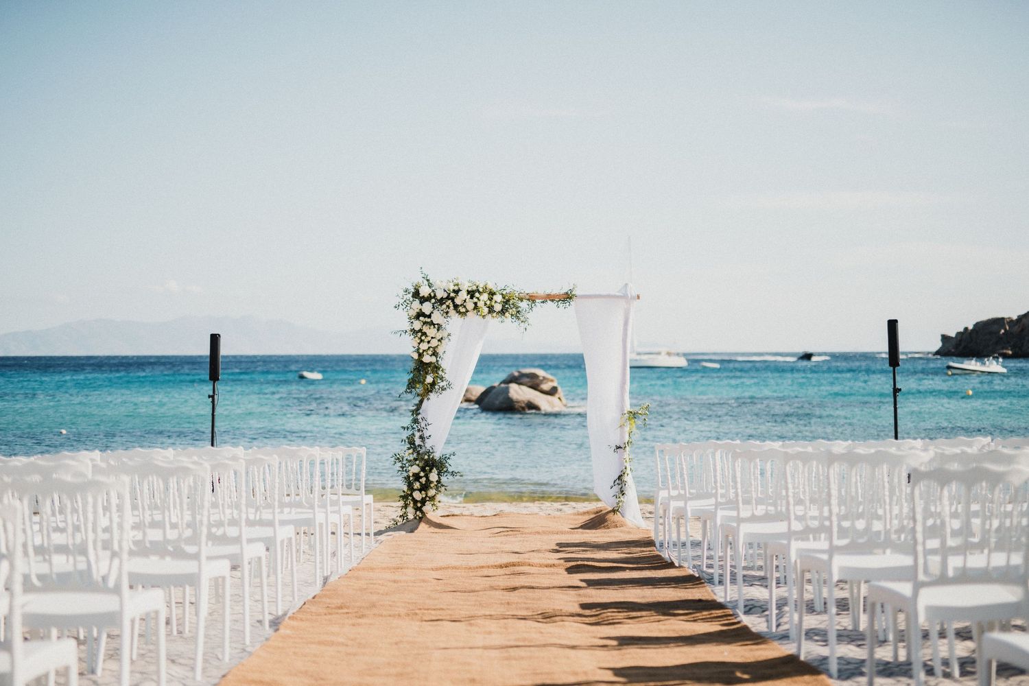 White wedding ceremony setup with chairs and floral arch on wooden boardwalk overlooking ocean.