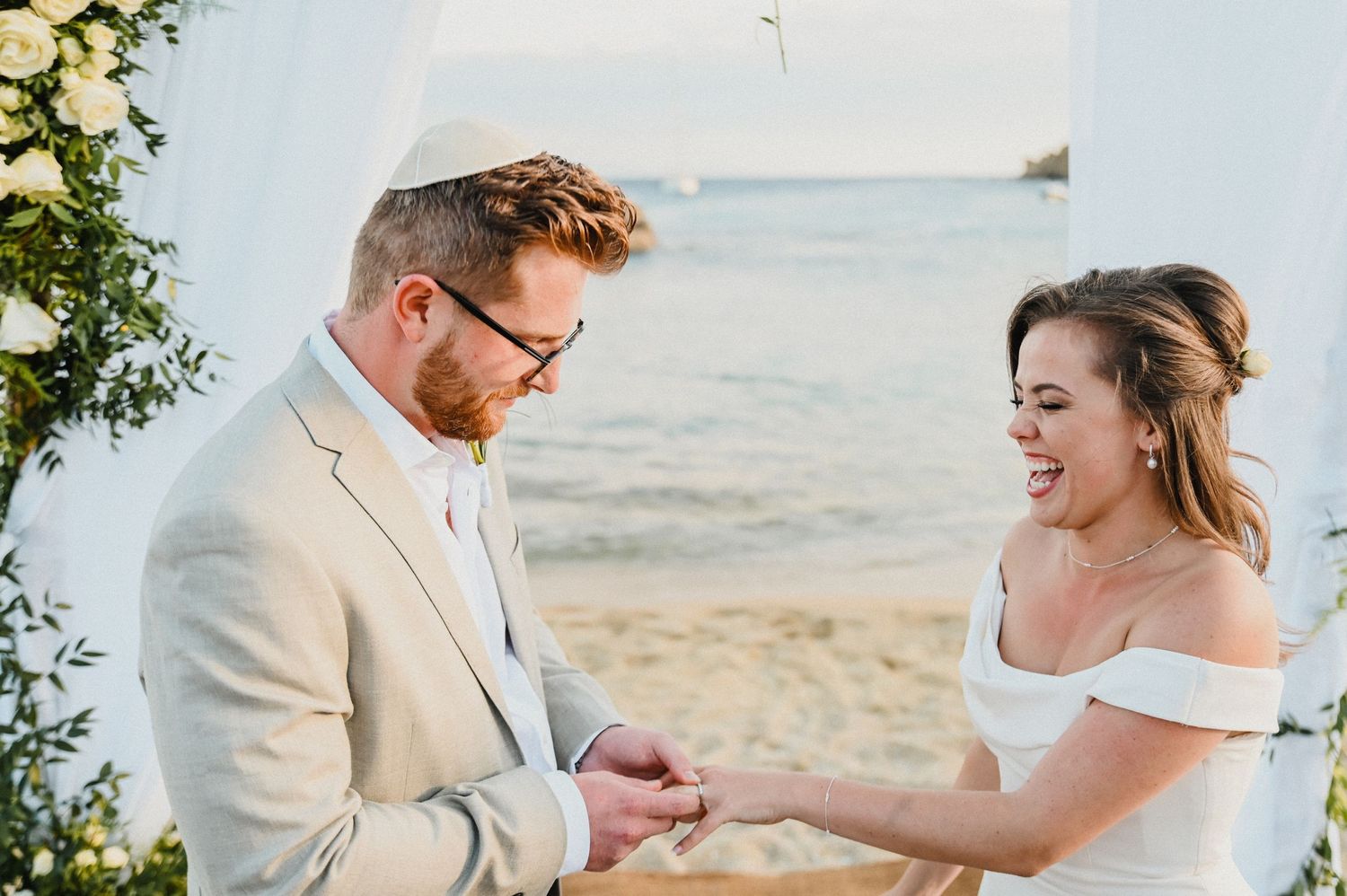 Couple exchanges rings during beach wedding ceremony beneath white fabric and floral arch.