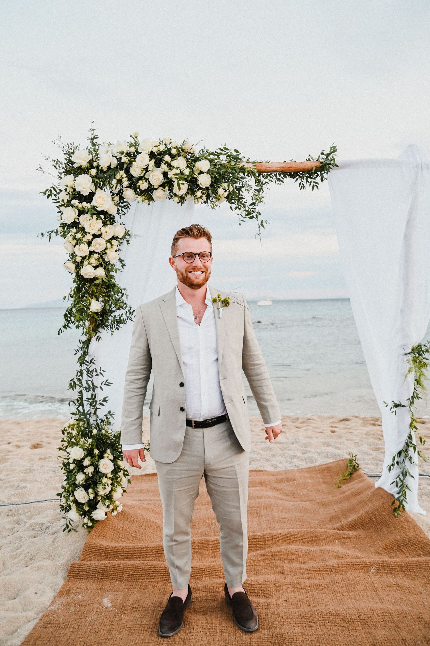 A beachside wedding ceremony setup featuring an elegant floral arch with white roses and greenery.
