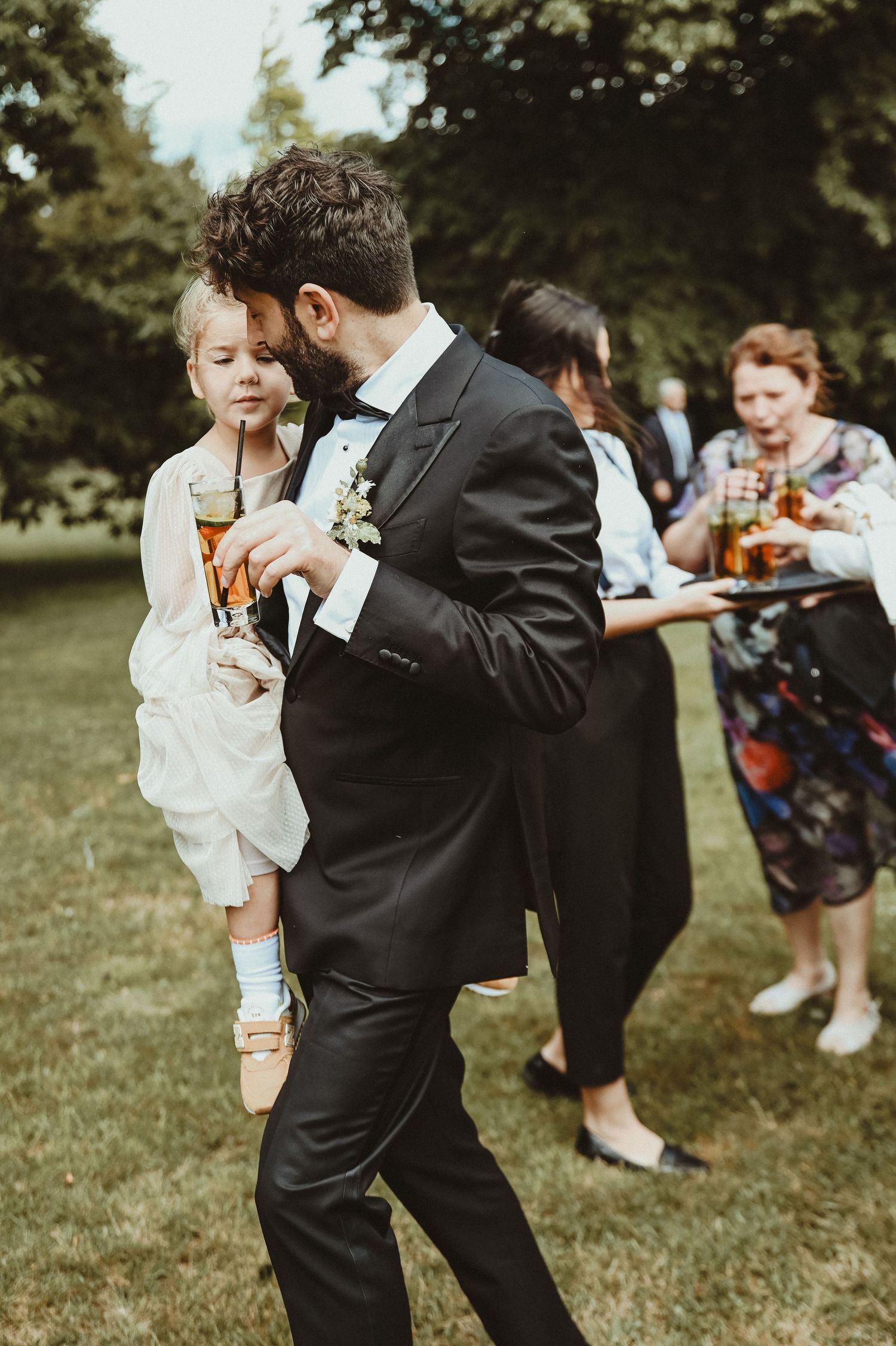A formal wedding guest dances while holding a small child at an outdoor reception.
