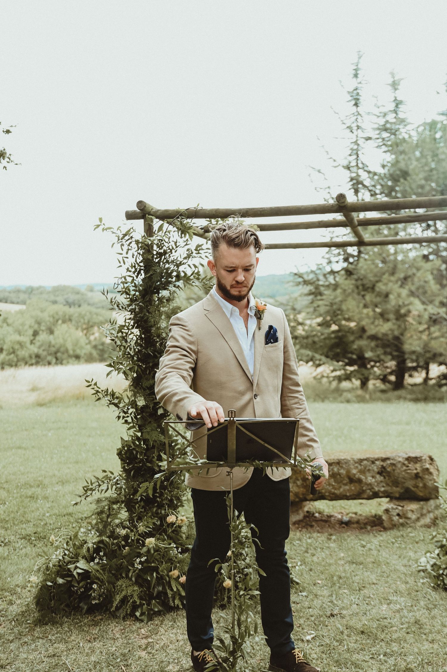 A groom in a beige suit jacket holds a box while standing under a rustic wooden arch decorated with greenery.