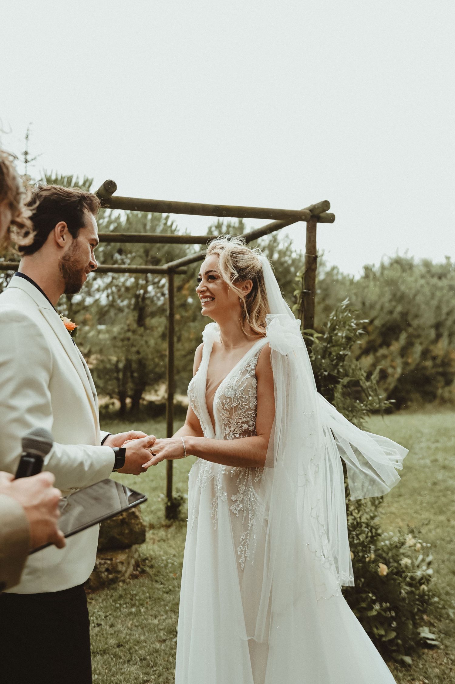 A couple exchanges vows during their wedding ceremony beneath a rustic wooden arch in a garden setting.