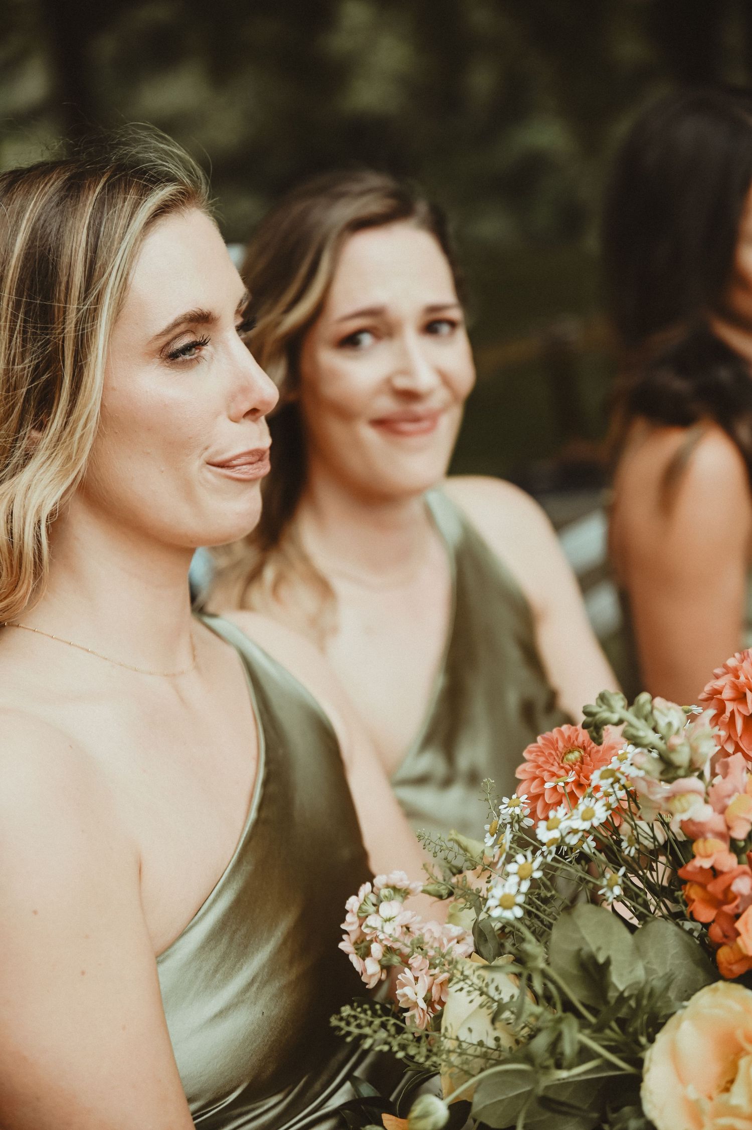 Bridesmaids in sage green dresses hold coral-colored floral bouquets at an outdoor wedding.