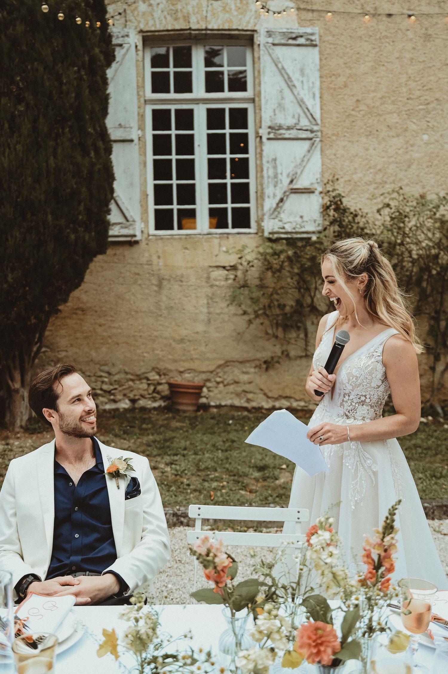Outdoor wedding reception speech with rustic stone building and floral table decorations.