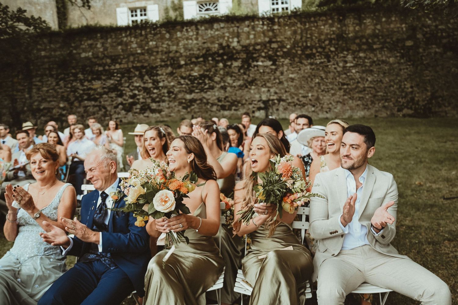 Wedding guests seated in rows hold bouquets while watching an outdoor ceremony against a stone wall.