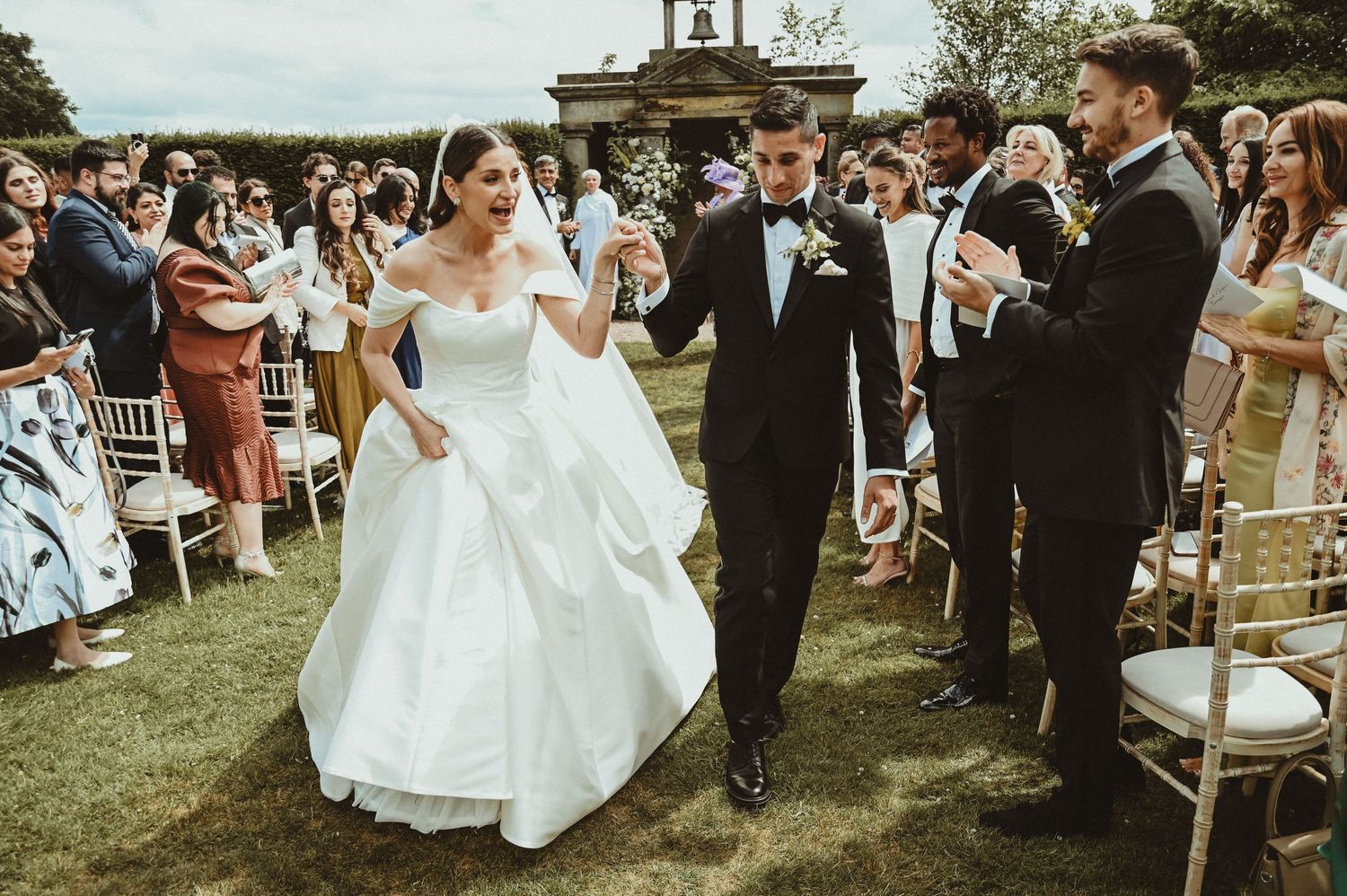 Newlyweds walking down the aisle while guests celebrate with confetti at an outdoor ceremony.